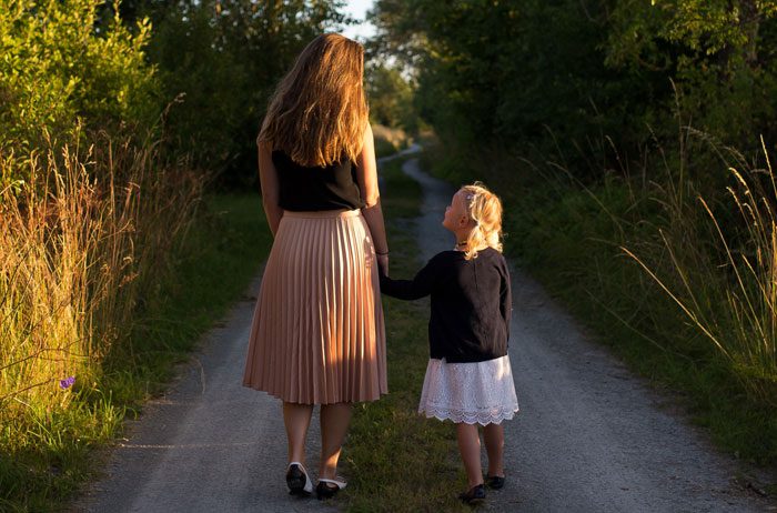 A mother/daughter pair walking down a nature trail between trees.